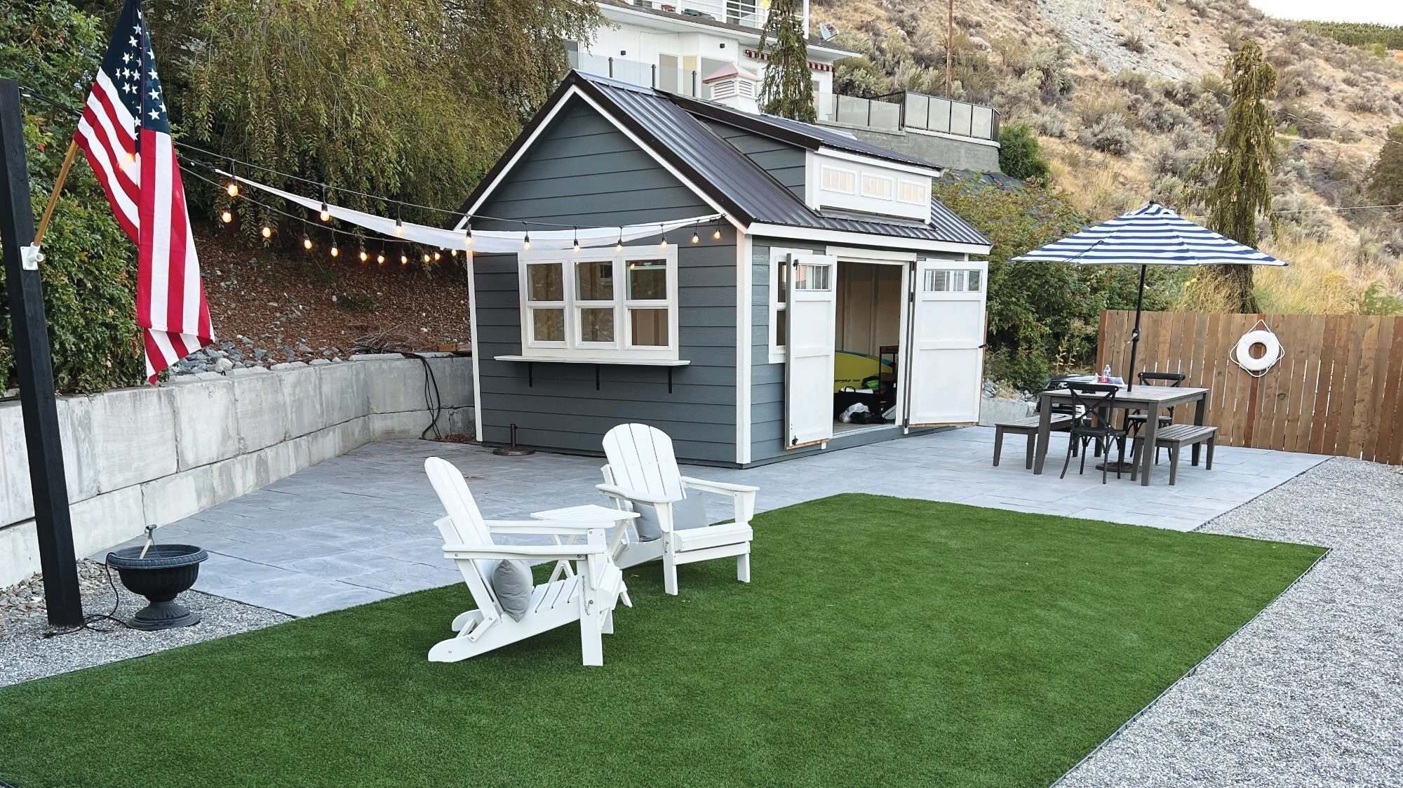 CABANA_IMG_1001 copy-16-9-2000x1125-min In a hilly backyard setting, this small grey shed with white trim features string lights and two white chairs on artificial grass. An outdoor table with a striped umbrella accompanies an American flag, capturing the charm of second homes in regions where Washington is a happy hunting ground for waterfront lots.