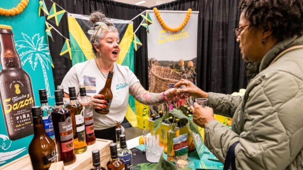 A vendor with tattoos offers a customer a drink sample at a booth displaying various liquor bottles and Caribbean-themed decor.