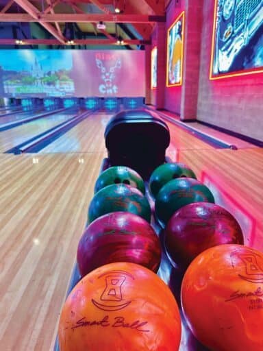 A vibrant bowling alley showcases six colorful balls on a rack, set against the backdrop of neon lights and artistic lane graphics. This scene could be straight out of a film festival held in March and April in Seattle, where art meets everyday fun.