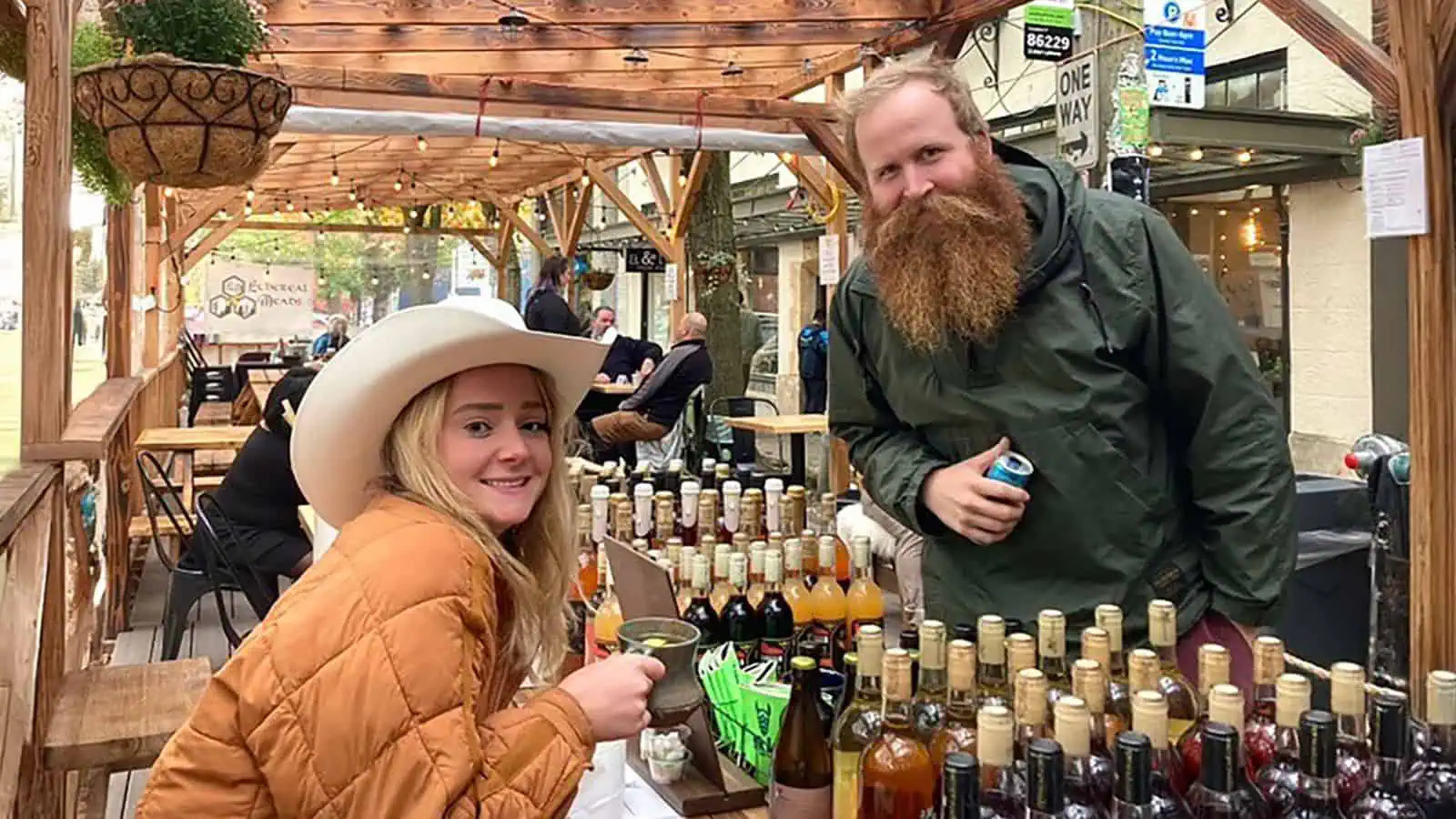 Amidst the festive outdoor scene, a woman in a cowboy hat and a bearded man stand at a table with various bottles, crafting their winter drinking plan. In the background, others enjoy the crisp Seattle air while seated at nearby tables.