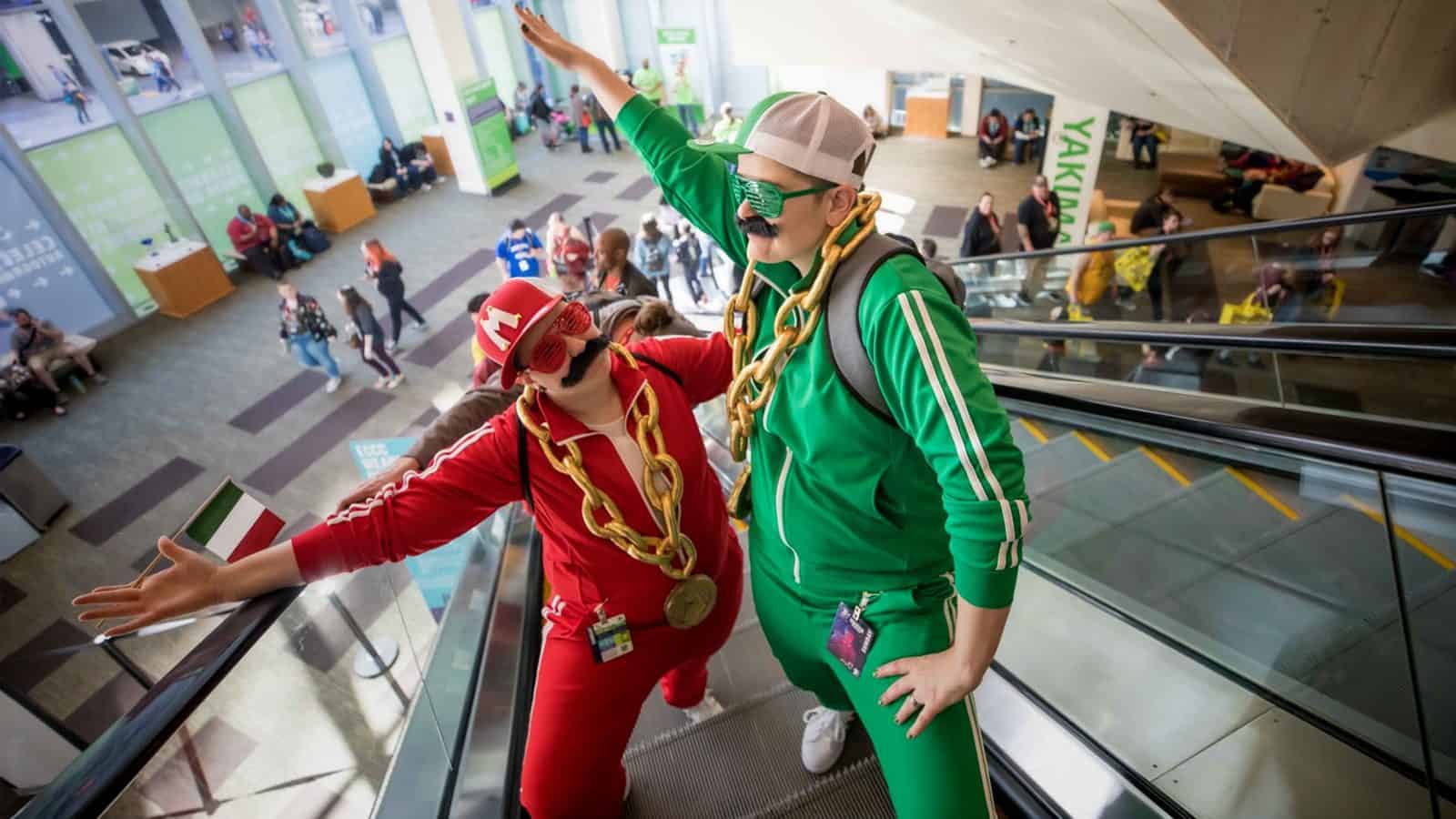 SuperMarioCosplay-EmeraldCityComicCon_1600x900 Two people in red and green tracksuits, with fake mustaches and oversized chain necklaces, pose on an escalator at a bustling event venue, capturing the eccentric spirit of Nerd Capital, America.