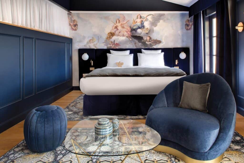 Luxurious hotel room with a double bed, elegant navy decor, and a mural above the bed invites guests to indulge. Relax in the plush chair or enjoy wines from Southern France at the glass coffee table, surrounded by a patterned rug on the wooden floor. Perfect for a charcuterie evening.