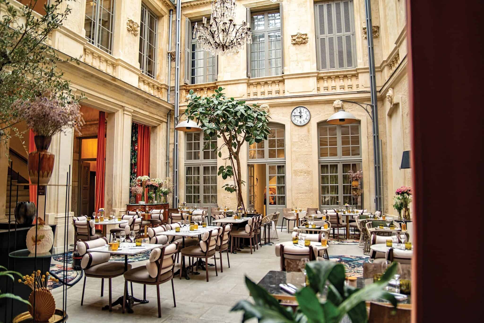 Chic indoor courtyard restaurant with elegant decor, multiple tables set for dining, a large clock on the wall, and charcuterie boards paired with fine wines. Natural light streams through tall windows, creating the perfect ambiance for a wine party in the Languedoc region.