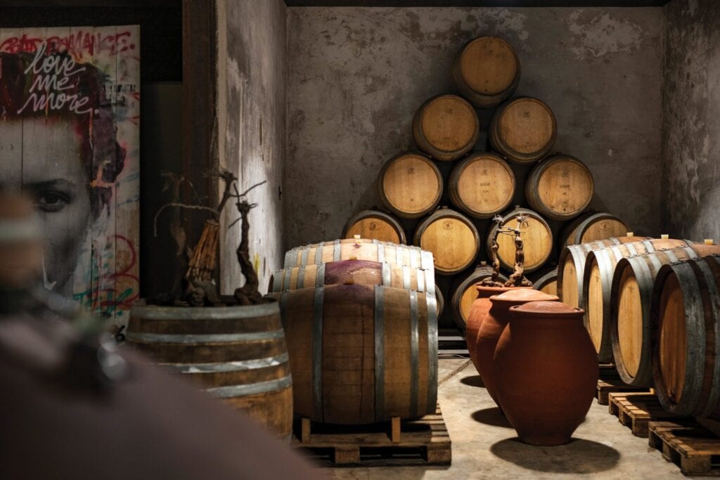 A wine cellar with stacked wooden barrels forming a pyramid and large clay pots sets the stage for a charming charcuterie display. In the background, a mural of a person's face whispers "love me more," inviting you to imagine a wine party in the Languedoc region.