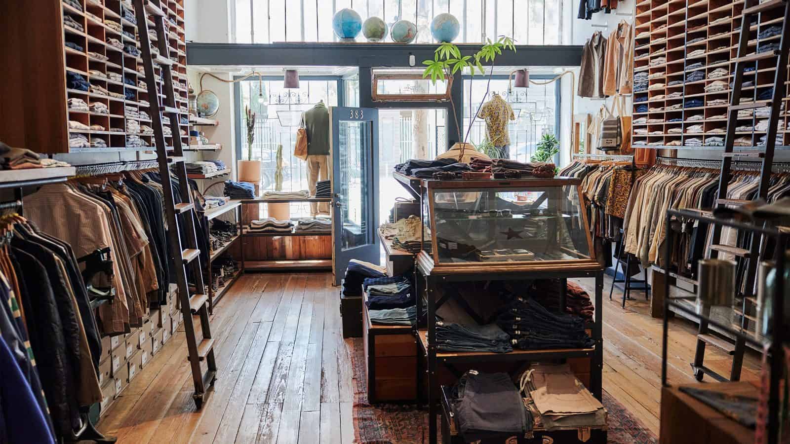 A Seattle clothing store interior with wooden floors, shelves filled with folded clothes, and racks of hanging garments. A Taylor Stitch glass display case is in the center, and globes adorn top shelves.