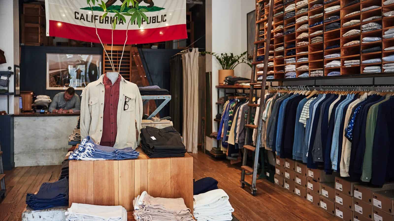 TaylorStitch-FirstSeattleStore_1600x900 The interior of the first Taylor Stitch store in Seattle features folded pants and a shirt on a mannequin in the foreground. Shirts and jackets hang elegantly on racks, while a large California Republic flag proudly waves overhead.