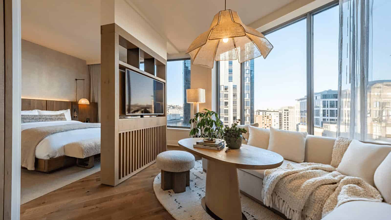 This modern hotel room, part of the exciting 2025 new hotels launch, features a connected bedroom and living area. Large windows, light wood furnishings, a round table with a plant, and a beige sectional sofa create an inviting space.