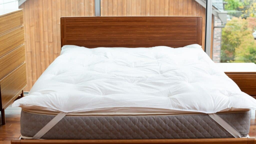 A neatly made bed with a white comforter is featured in the bedroom, highlighting the elegance of Bedrooms & More. A large window in the background allows natural light to illuminate the space, showcasing the quality of fine bedding.