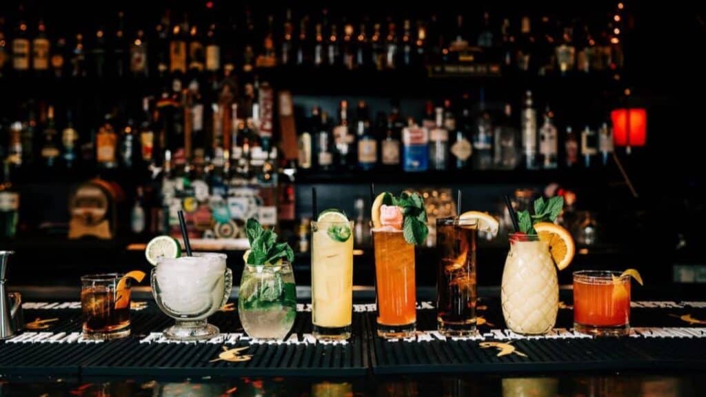 A line of colorful cocktails and mixed drinks garnished with fruits and herbs, displayed on a bar counter with a variety of bottles in the background.