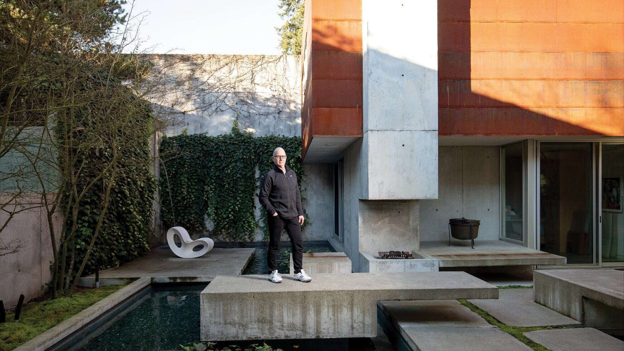 COBB_250124_sm_eric-cobb_3862_p copy-16-9-2000x1125-min A person stands on a concrete platform in a modern architectural courtyard, designed by architect Eric Cobb, featuring a pool, vine-covered walls, and a distinctive chair. This serene space exemplifies the allure of a meticulously crafted second home.