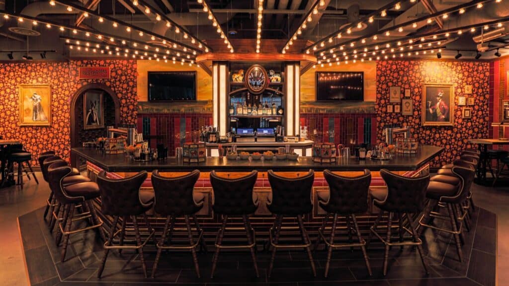 Image of an empty, ornate bar with a U-shaped counter, lined with high-backed stools, warm lighting, patterned walls, and framed art.