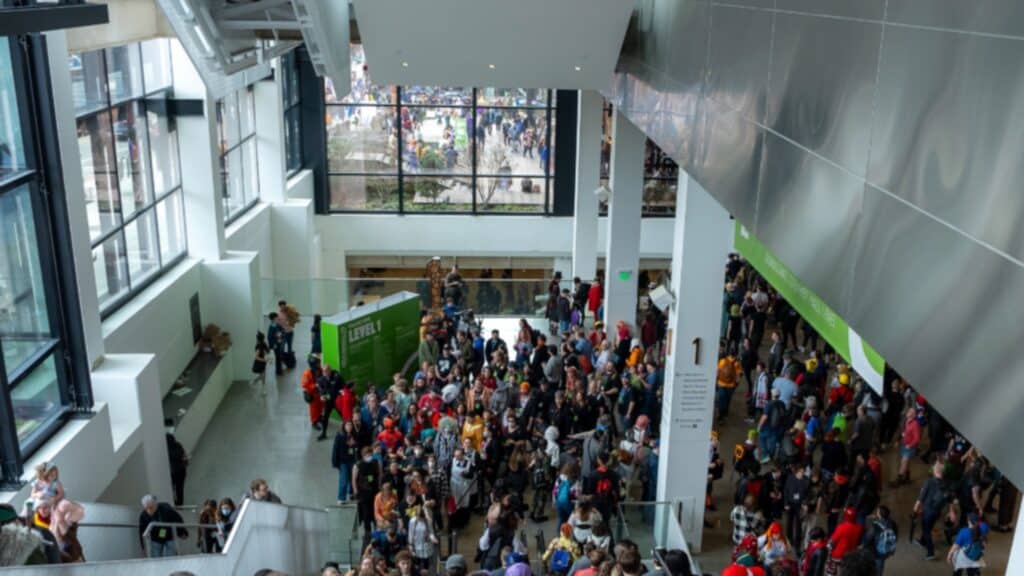 A large crowd of people gathers in a spacious, modern indoor area with high windows and signs, reminiscent of the largest Comic Con event, viewed from above on a lively weekend.