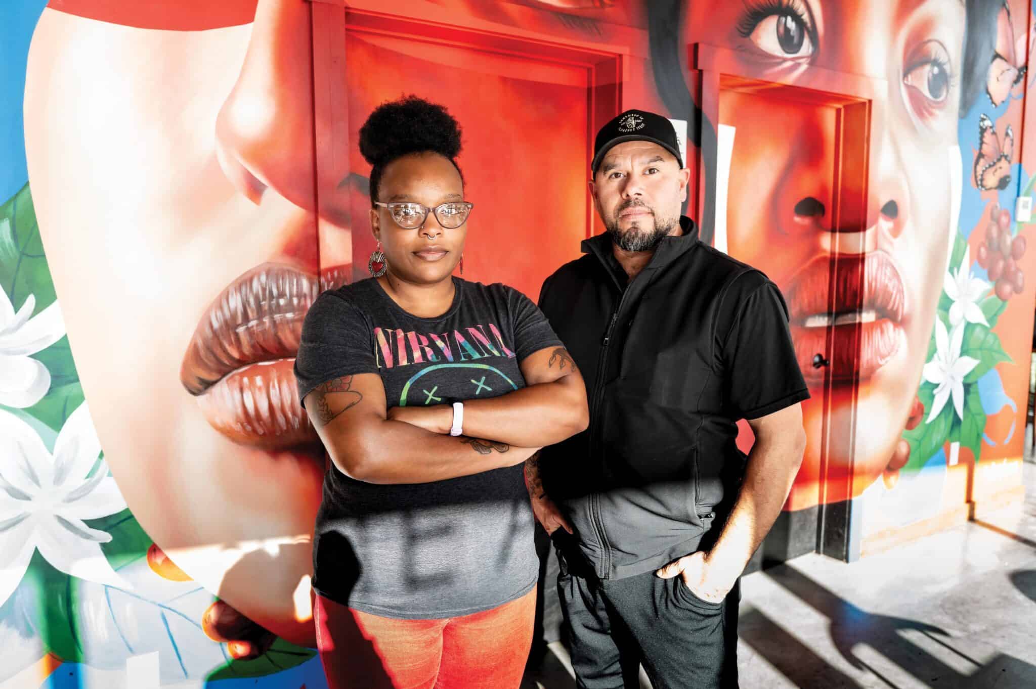 Two people stand in front of a colorful mural featuring large human faces and flowers, their expressions suggesting a budding romance.