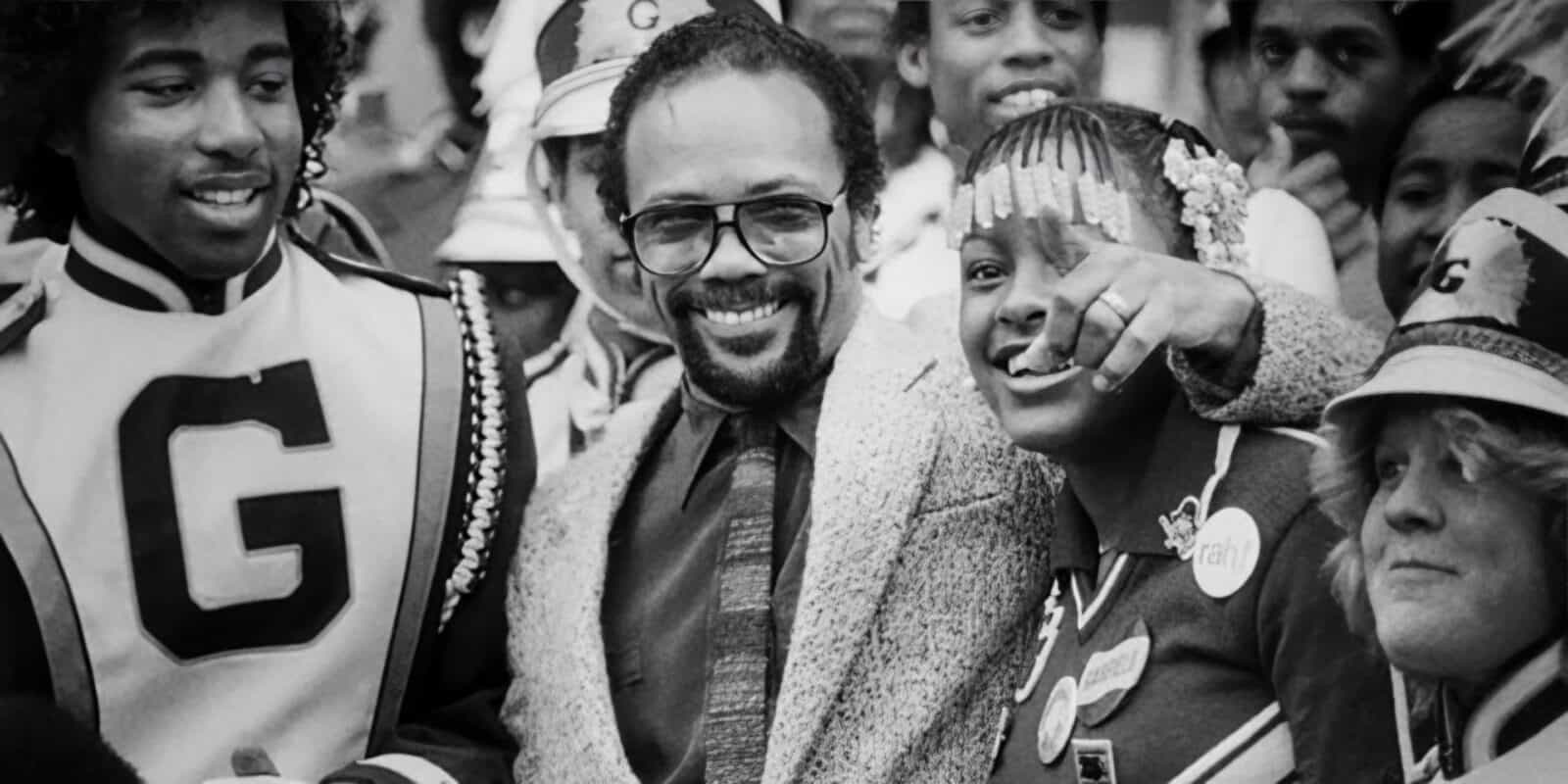 GarfielfJazz-TributeToQuincyJones_1600x900 A lively group gathers, some in marching band uniforms, smiling and looking around. At the center stands a man in glasses and a suit, embodying Quincy's legacy, surrounded by others in festive attire.