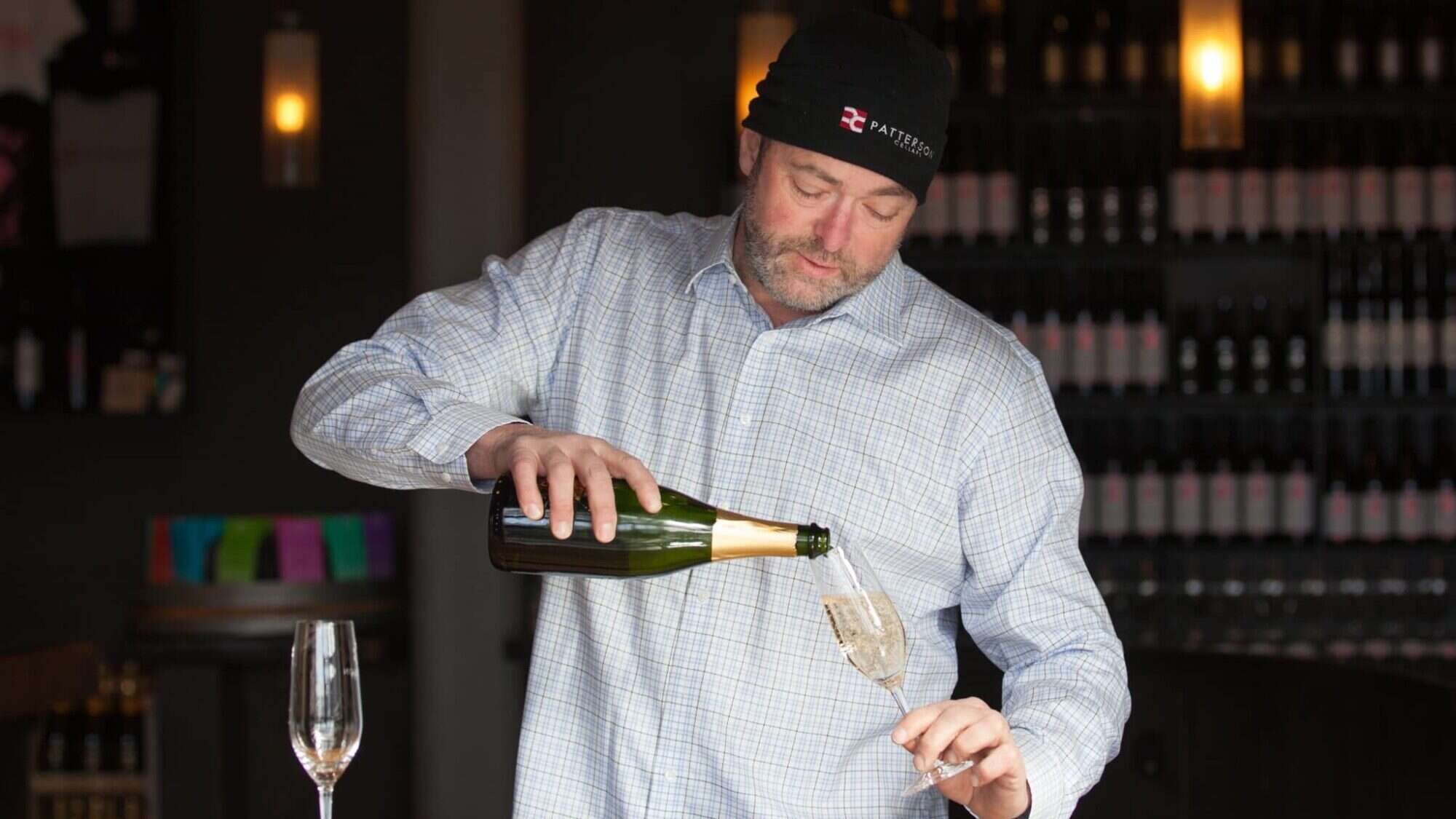 Man in a checkered shirt and beanie pouring champagne into a glass at a bar with wine bottles in the background.