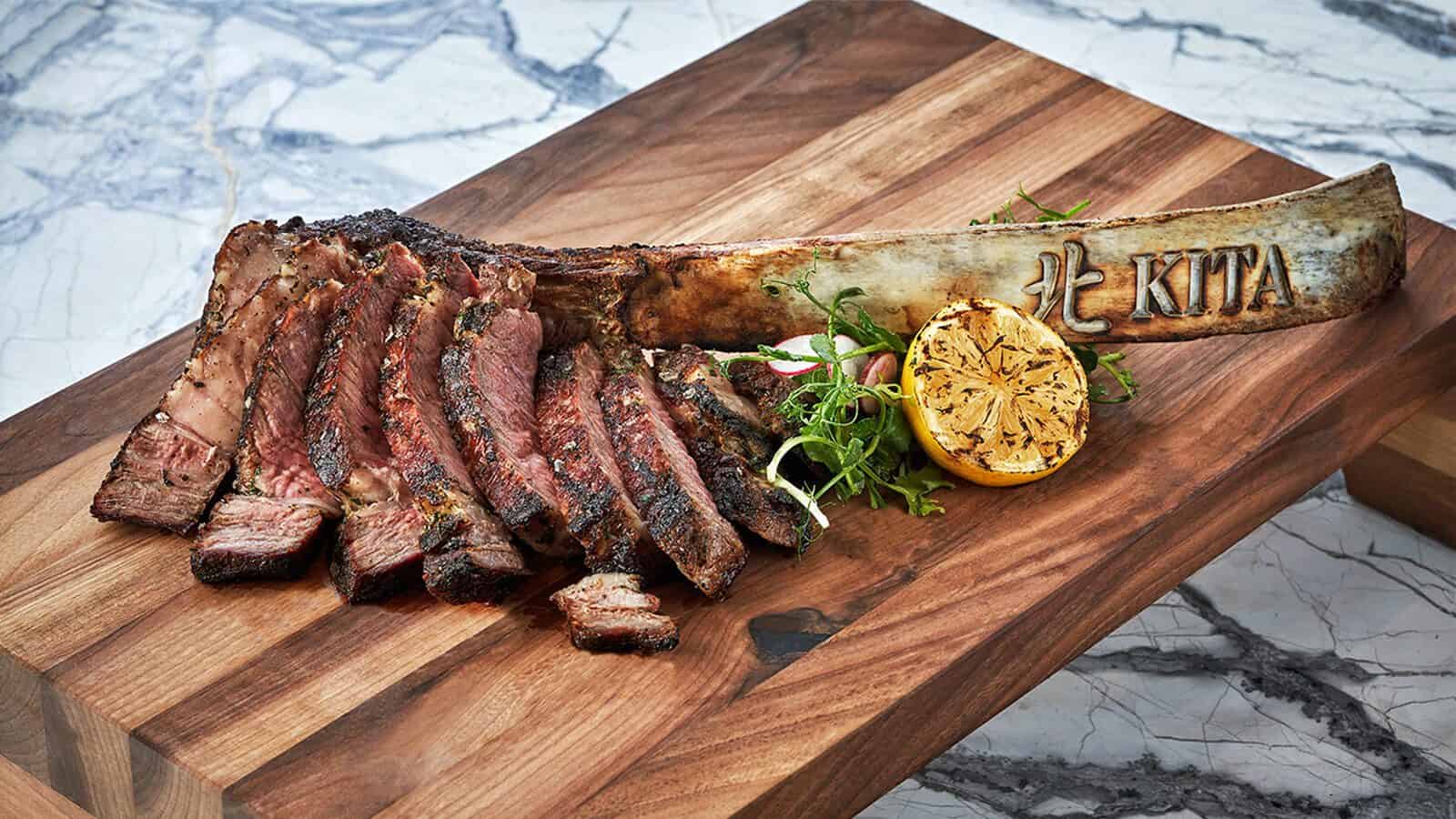 Sliced grilled tomahawk steak on a wooden board with a branded bone, garnished with grilled lemon and greens—a perfect dish to enjoy after a day skied in Park City.