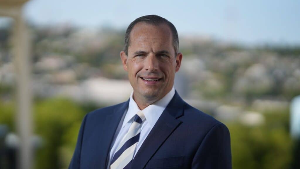 A real estate broker in a suit and striped tie stands outdoors with a blurred landscape in the background. He is smiling and facing the camera, ready to discuss housing prices with confidence.