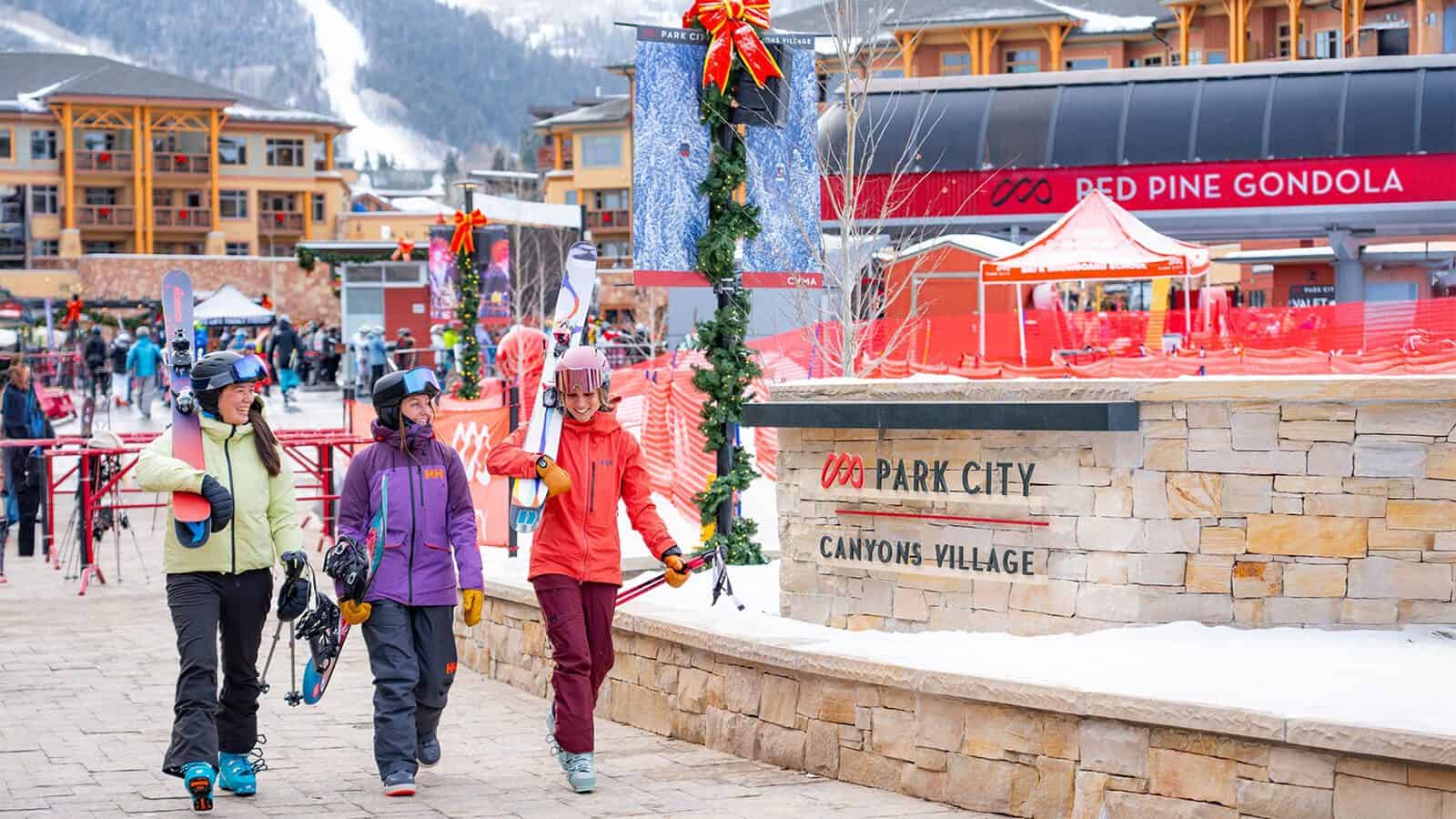 Three people carrying skis stroll past a sign reading "Park City Canyons Village," with a ski lift and buildings in the backdrop. It's the perfect scene for a weekend in Park City, boasting some of the best snow for those eager to have skied these renowned slopes.