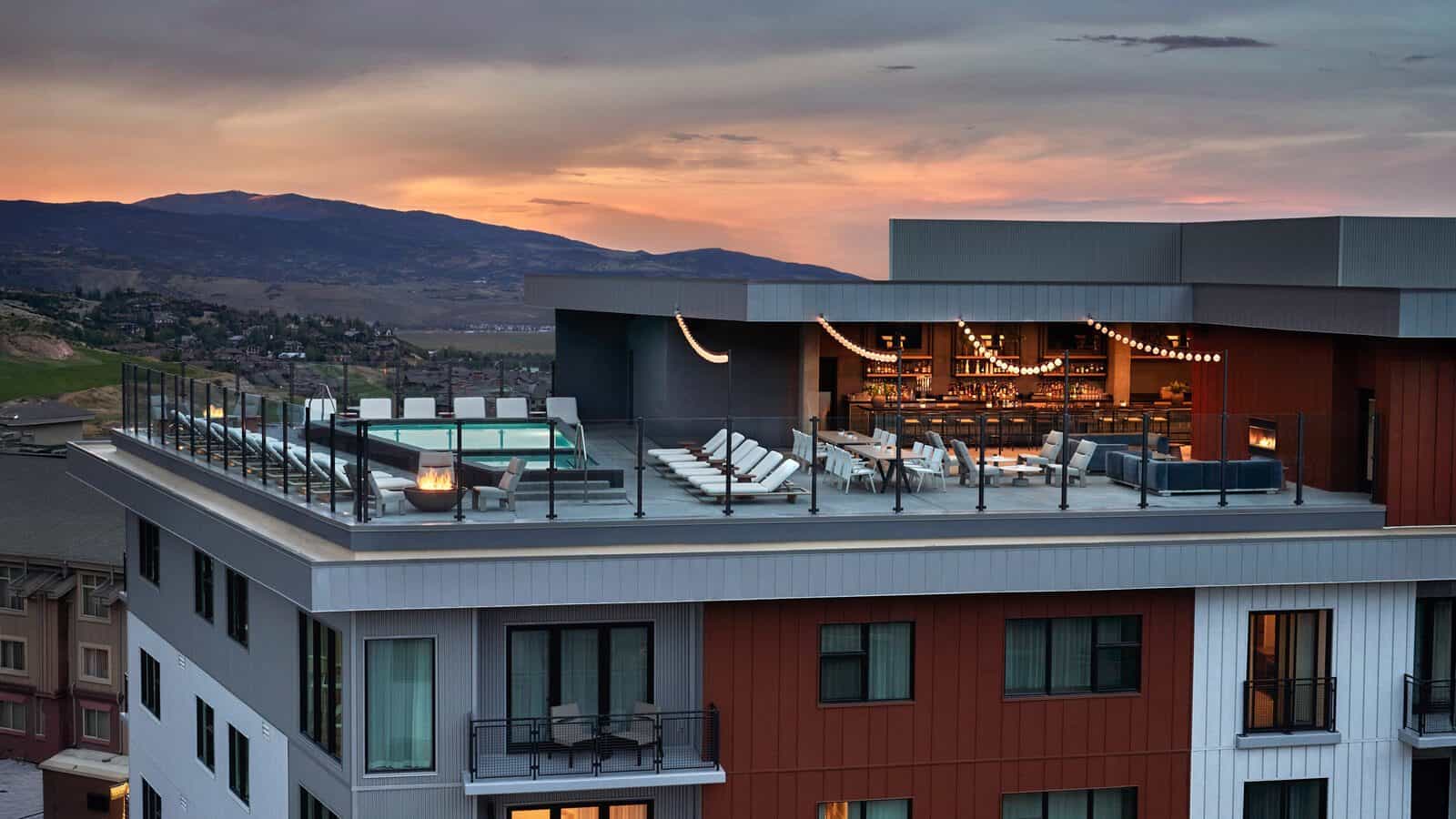 A rooftop lounge with a pool, seating area, bar, and string lights basking in the glow of a mountainous sunset backdrop is the perfect way to unwind after the best snow in Park City on a weekend escape.