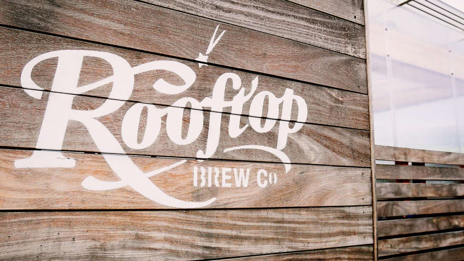 RooftopBrewCo-Signage_1600x900 A sign on wooden planks boasts the logo "Rooftop Brew Co" with a stylized font and an arrow motif, celebrating the maritime tradition of brewing.
