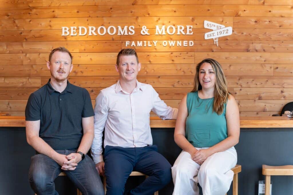 Three people sit on stools in front of a wooden wall adorned with the "Bedrooms & More Family Owned" sign. The two men and woman smile at the camera, showcasing their passion for quality bedrooms and fine furniture.