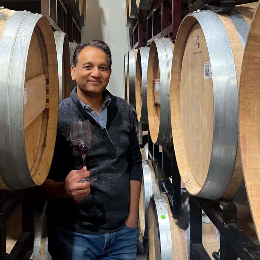 In celebration of 25 years, a person holds a glass of red wine, standing among wooden barrels in the cellar, donning a dark sweater—a perfect embodiment of the Taste Washington spirit.