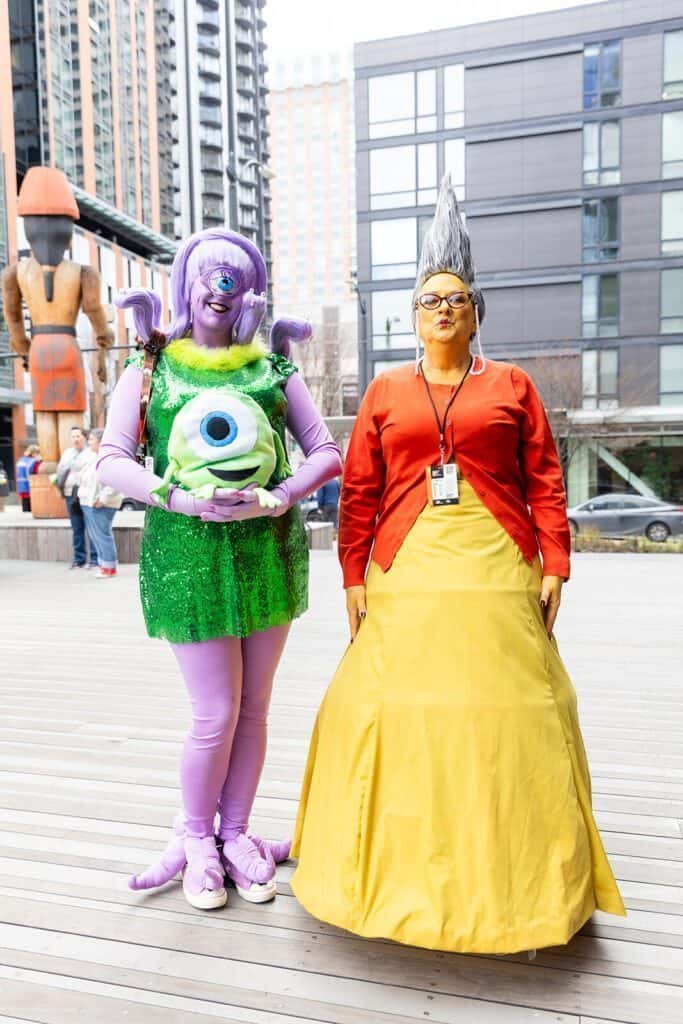 Two people in colorful costumes stand outdoors. One is in a green and purple outfit with a single eye on the chest, and the other is in a red top and yellow skirt with a tall gray wig.