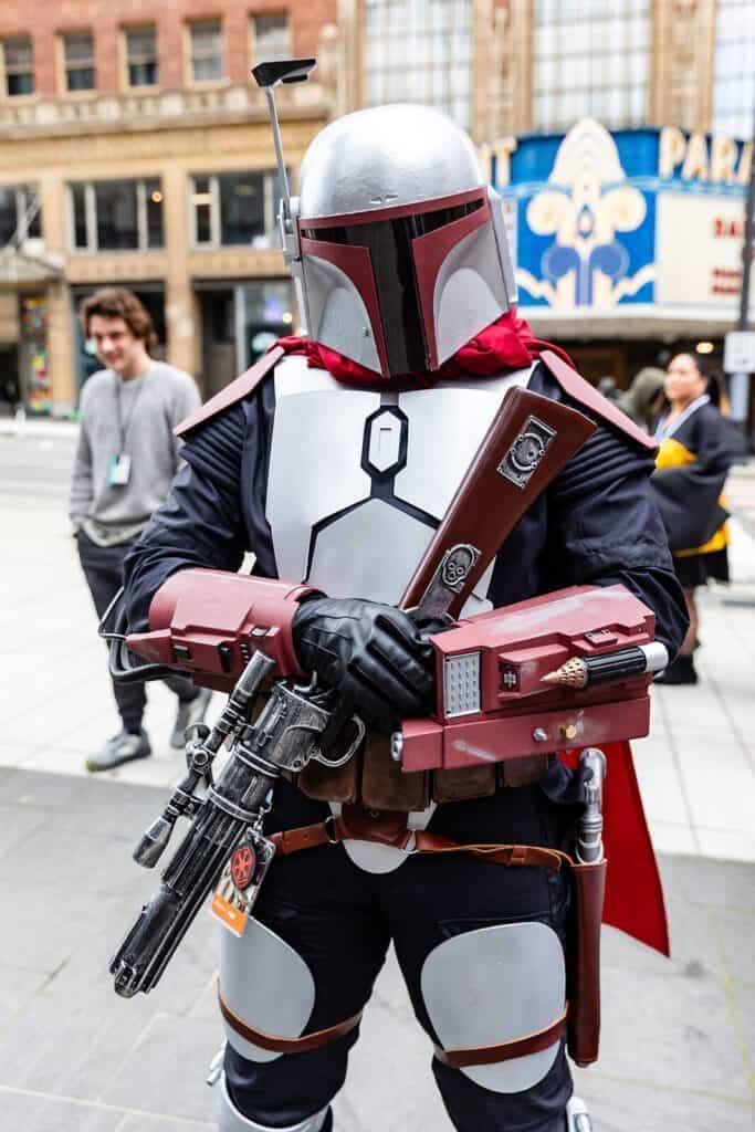 Person in detailed Mandalorian armor cosplay standing in an urban setting, holding a prop blaster.