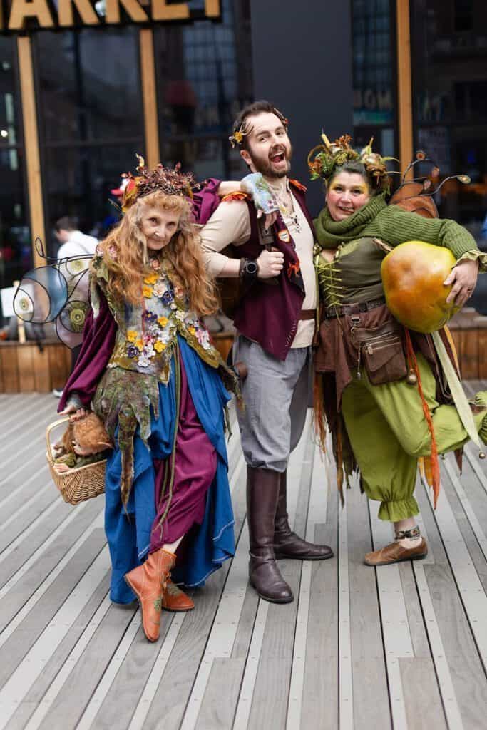 Three people in colorful, fantasy-themed costumes with flowers and accessories pose enthusiastically on a wooden deck.