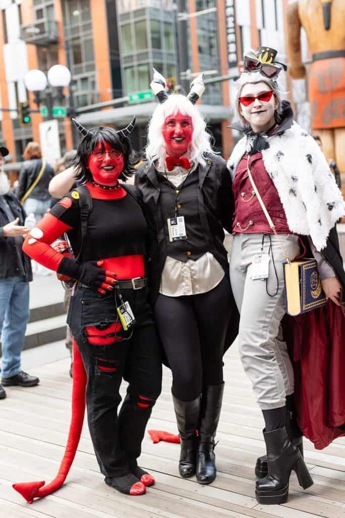 Three people in fantasy costumes pose for a photo on a boardwalk. Two wear red body paint, horns, and tails, while the third wears a cape and holds a book.