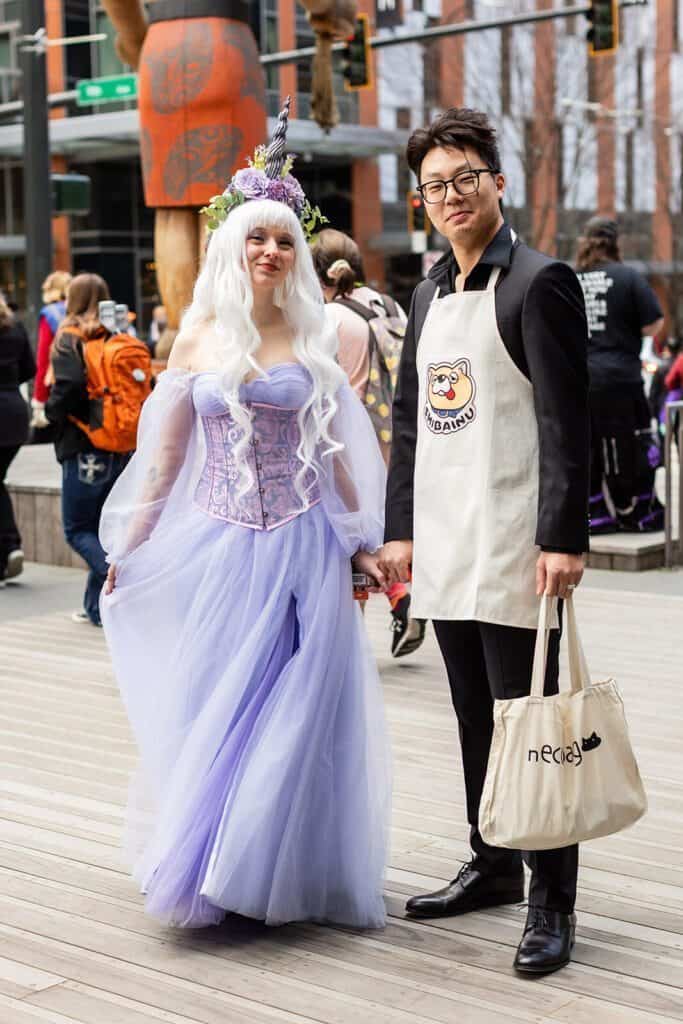 A woman in a purple dress and flower crown stands next to a man wearing glasses and a beige apron. They are outdoors on a wooden pathway with people and buildings in the background.