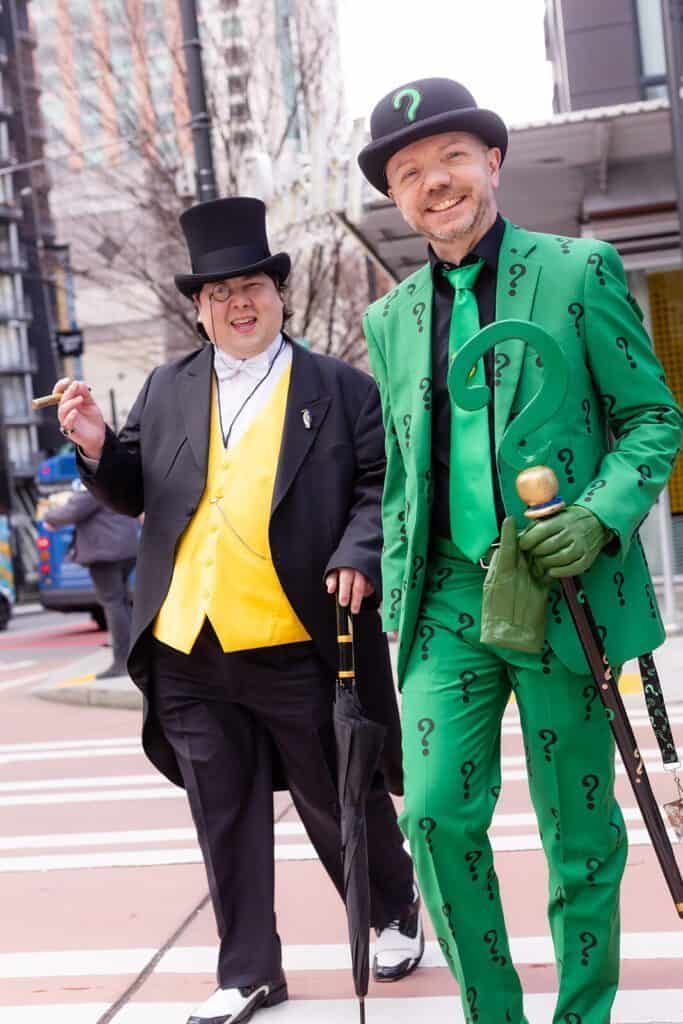 Two people in costume walking on a city street: one in a black top hat and yellow vest, the other in a green suit with question marks and a bowler hat.
