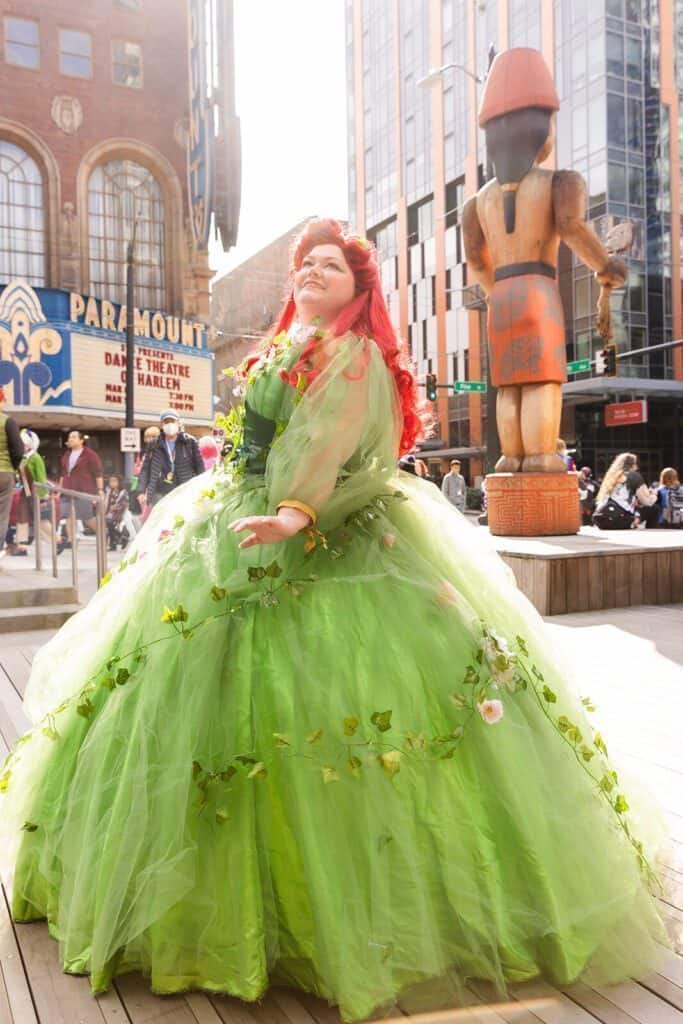 Person in an elaborate green gown decorated with flowers poses outdoors. A large wooden sculpture and a theater marquee are visible in the background.