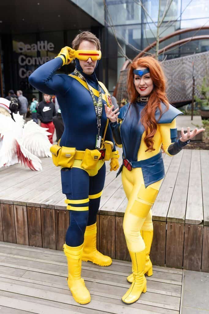 Two people in matching blue and yellow superhero costumes pose outdoors, with convention center signage in the background.