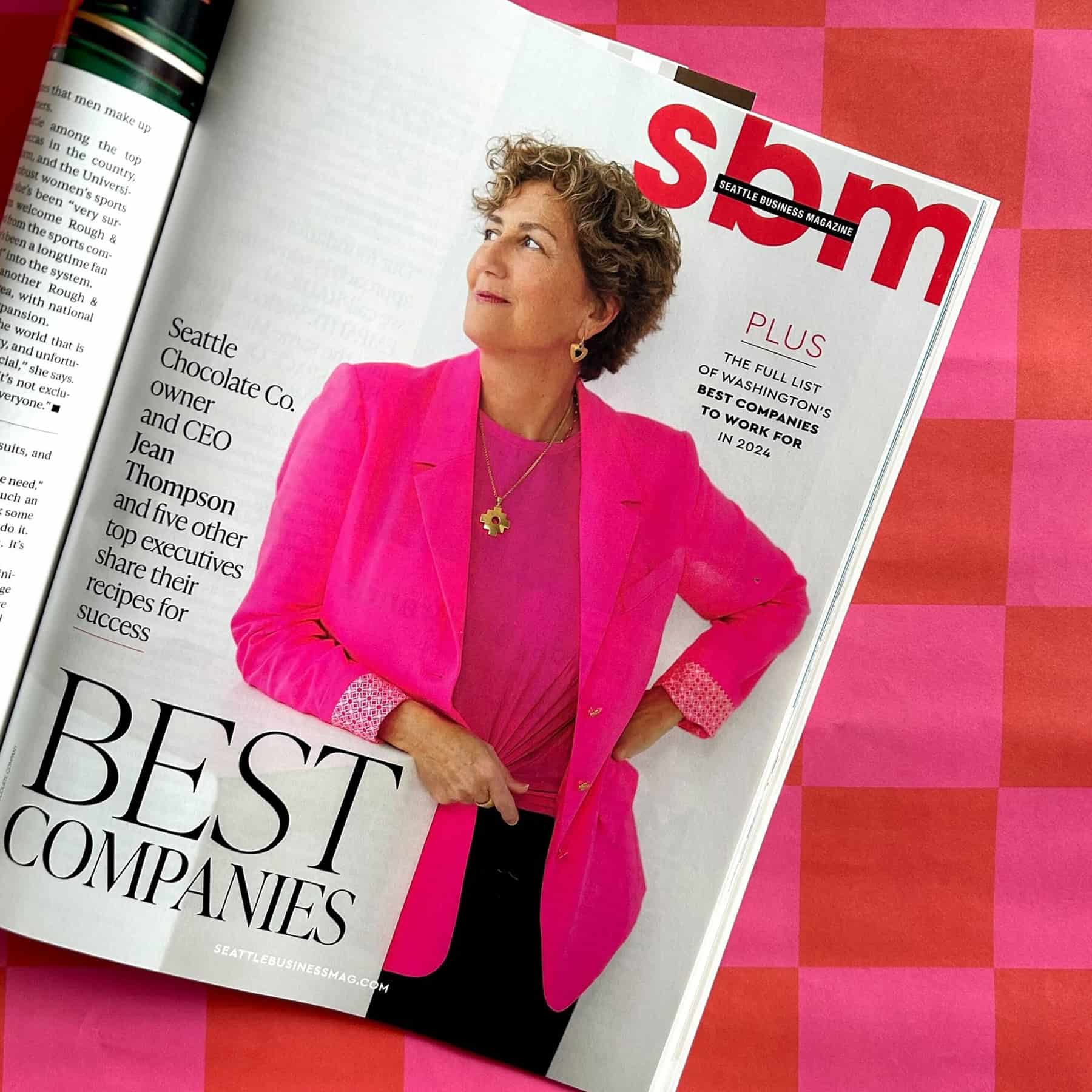 A magazine lies open to a page showcasing Seattle Chocolate Co. CEO Jean Thompson in a bright pink blazer. The headline reads "Best Companies," weaving the story of her remarkable leadership and the success of her brand.