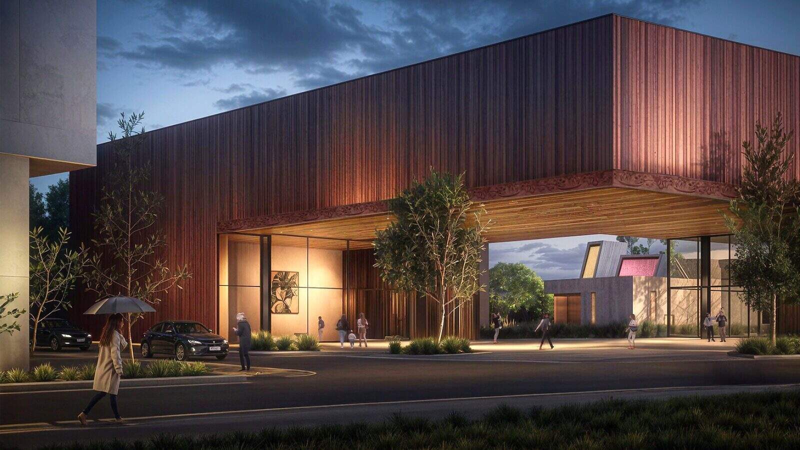 SeattleUniversityMuseum-Interior02_1600x900 At dusk, beneath a modern building with a wooden façade, people stroll near Seattle U's vibrant campus. A person shields themselves with an umbrella as cars are parked nearby, and trees line the road in this bustling scene.