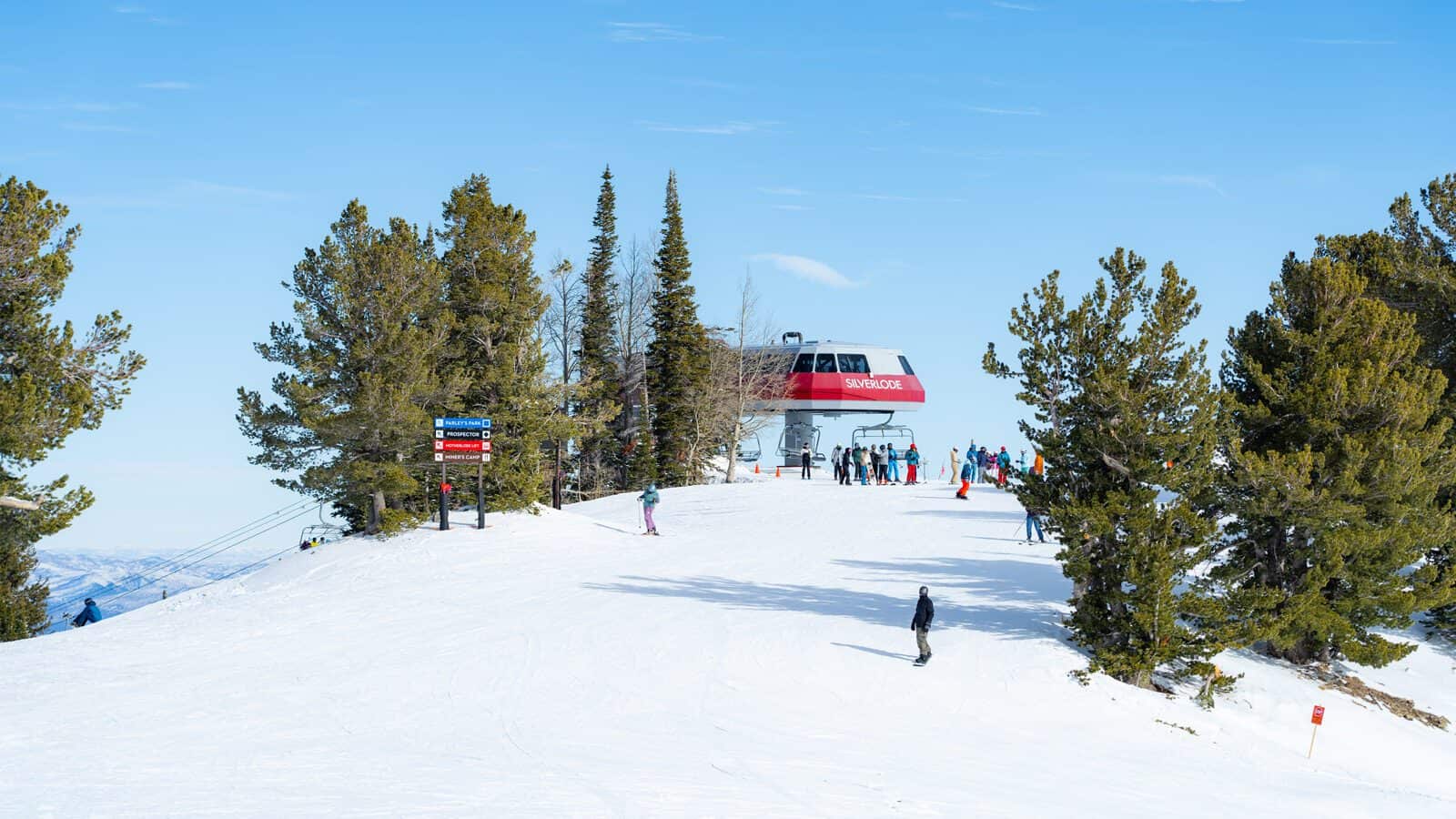 A group of skiers and snowboarders gather near a ski lift on the snowy slopes of Park City, surrounded by pine trees under a clear blue sky.