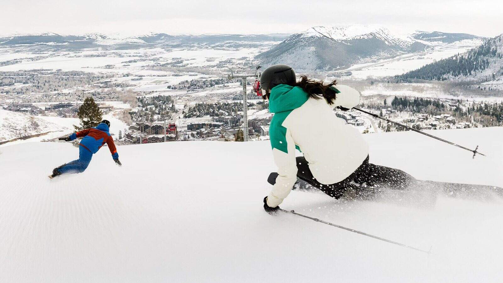 Skiing-ParkCityMountain_1600x900 Two skiers in colorful jackets expertly descend a snowy slope, part of the best snow in Park City, overlooking a mountainous landscape and a distant town.