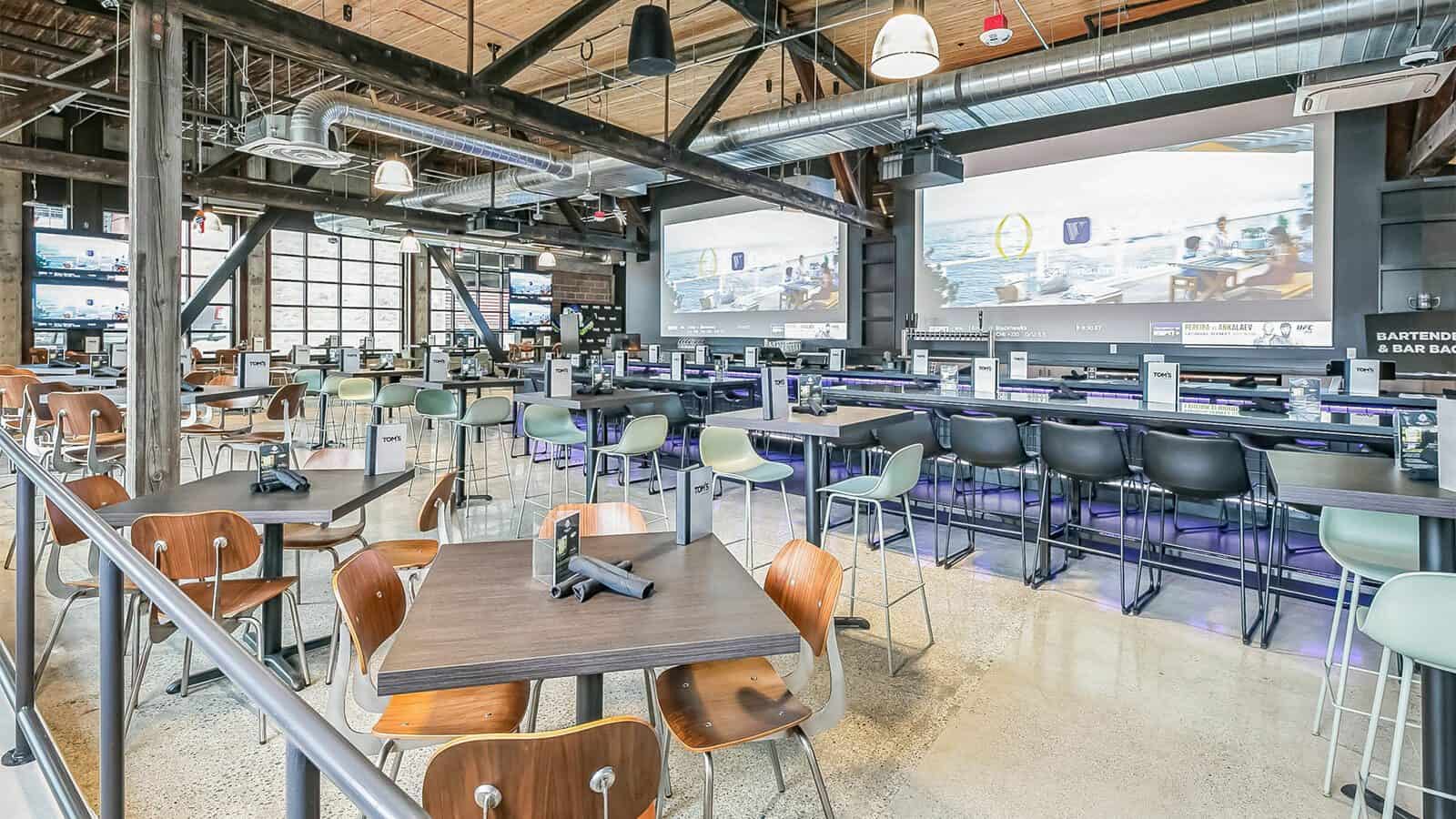 Located in the bustling heart of Uptown, Tom’s Watch Bar boasts a modern, spacious interior with high ceilings. Enjoy dining at tables and chairs perfectly arranged under large screens, all bathed in ample natural light.