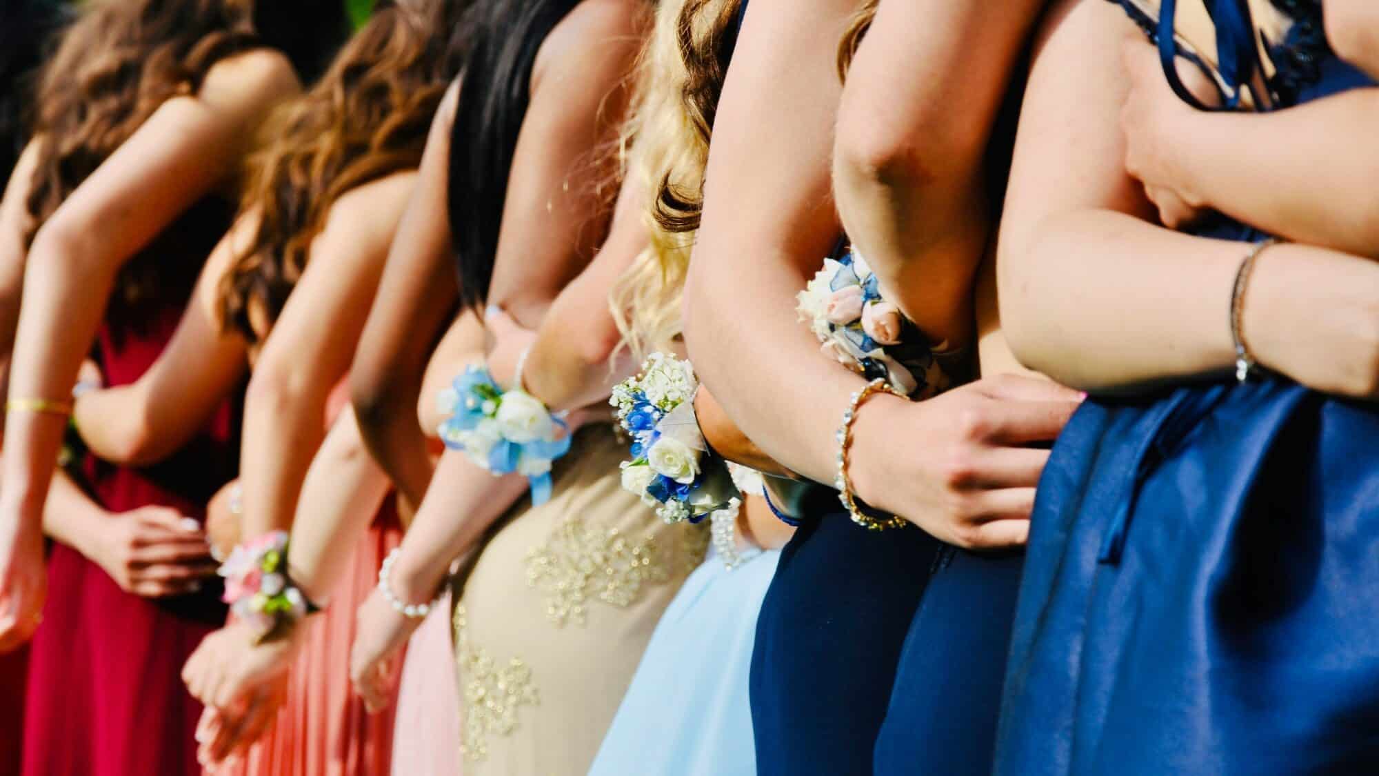 todd-cravens-7dCbl4BfoRc-unsplash-16-9-2000x1125 A line of people in formal dresses with corsages on their wrists, standing side by side and holding each other’s waists.