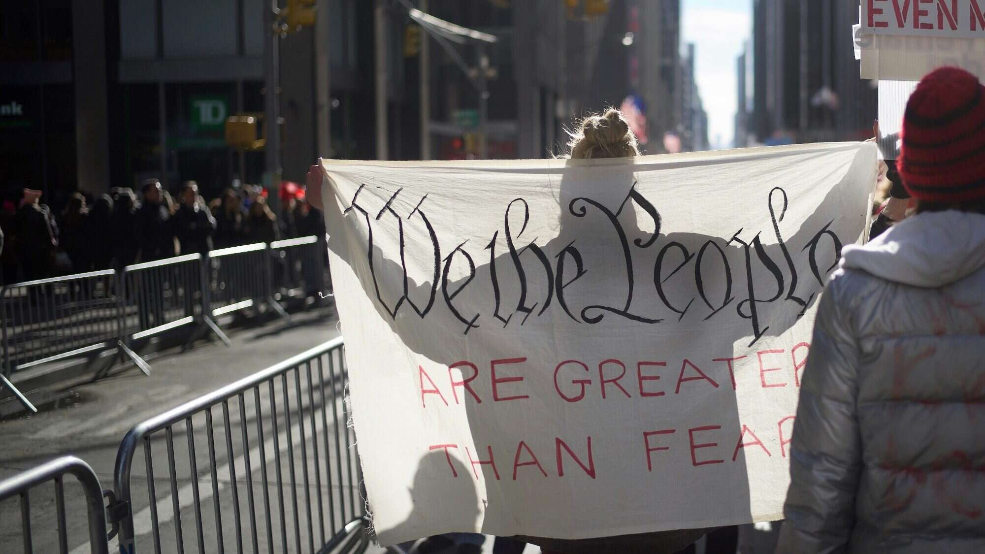 A person holds a sign reading "We the People Are Greater Than Fear" at a street demonstration, their voice resonating across all fifty states, with metal barricades and buildings in the background.