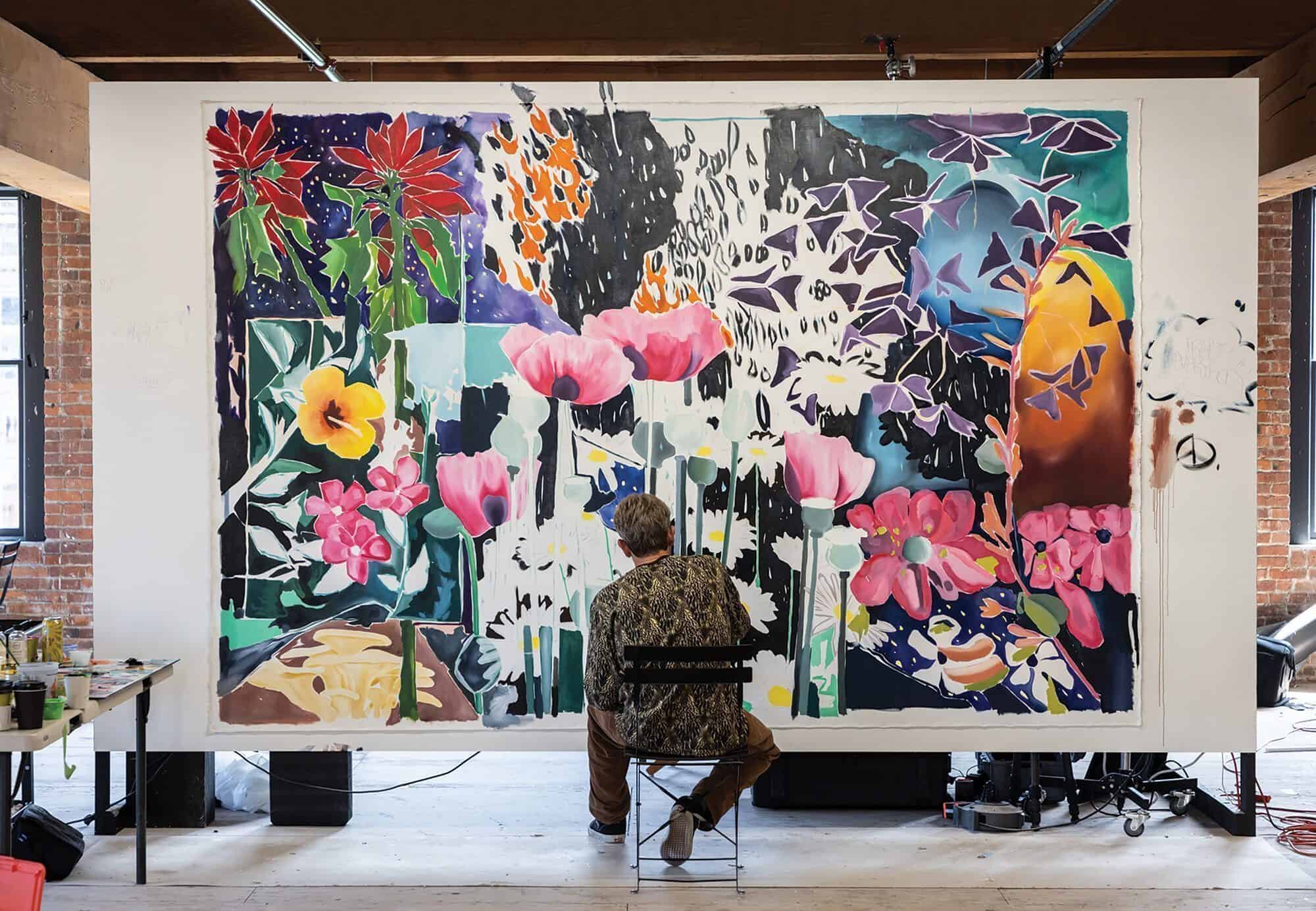 ART_HotelWestland_Art in Progress by Sean Barton copy A person sits on a chair painting a large, colorful floral mural on a white wall in a spacious, well-lit studio near Pioneer Square Seattle's historic Hotel Westland, blending creativity and local charm for ARTXIV.