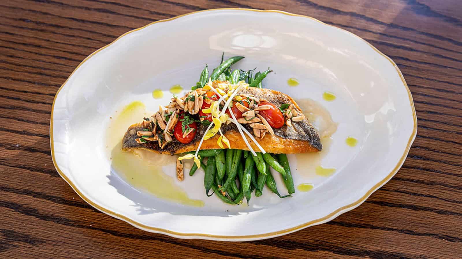 A cooked fish fillet topped with tomatoes, sliced almonds, and microgreens sits on green beans, drizzled with olive oil—inviting you to taste flavors as vibrant as A Stroll Through Old & New D.C., served elegantly on a white plate with a gold trim.