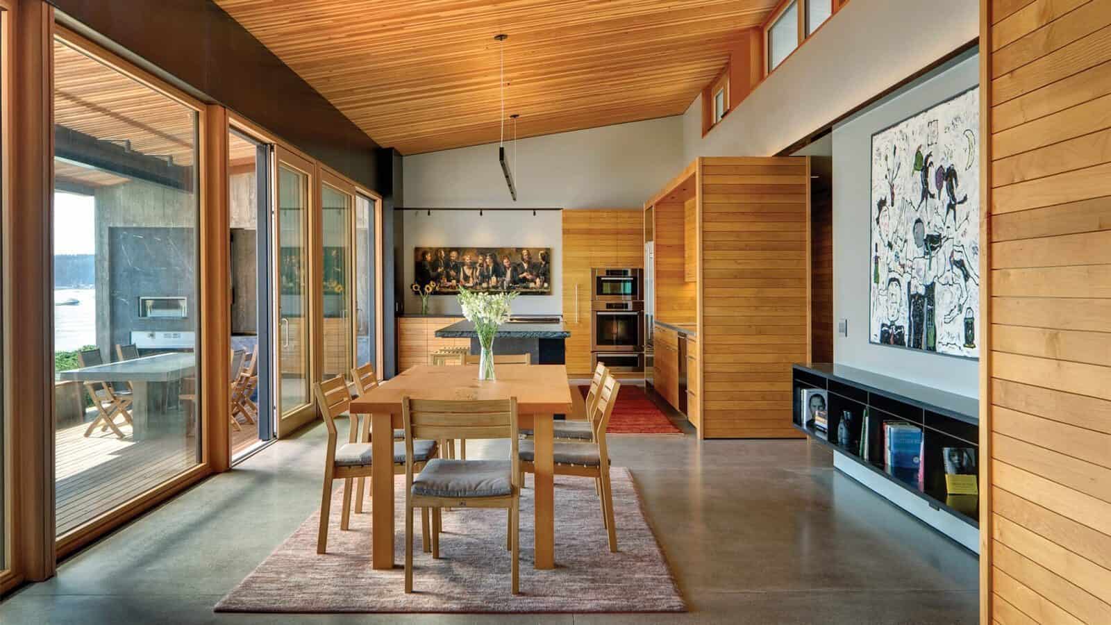 Modern dining area with wooden furniture, large windows, and an open kitchen; artwork hangs on the wall as natural light fills architect Eric Cobb's second home.