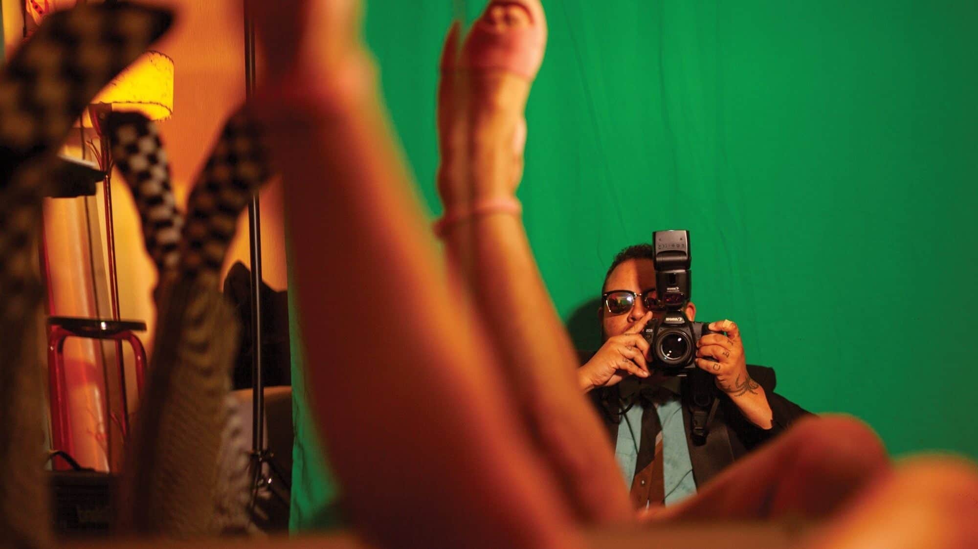 A person in sunglasses and a suit photographs someone lying down in front of a green screen, with the subject's legs visible—an image reminiscent of Seattle artist Fox Whitney’s bold explorations at On the Boards Seattle.