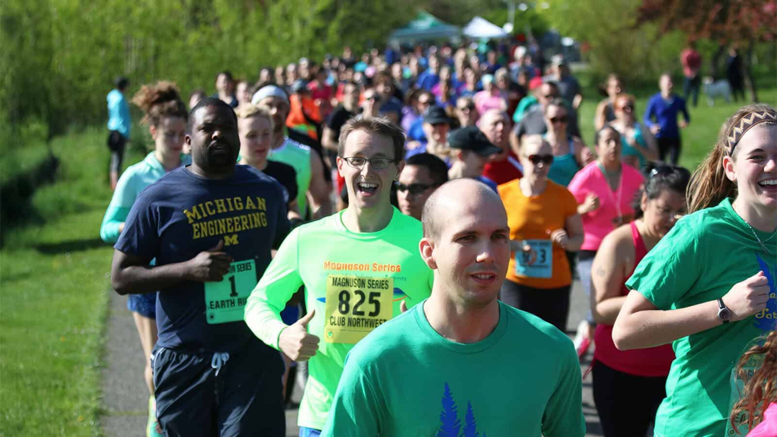 A group of runners participates in an outdoor race on a sunny day, celebrating Earth Day. Some wear colorful race bibs, and the surroundings include important elements like green grass and trees that highlight the ever-vibrant spirit of nature.