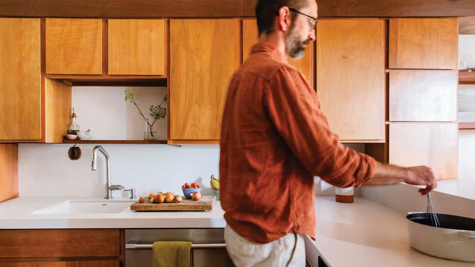 A person in an orange shirt stirs a pot on the stove in a kitchen with wooden cabinets reminiscent of Frank Lloyd Wright's style, while a cutting board with apples sits on the counter, evoking a sense of timeless elegance typically achieved through home renovations by architects.