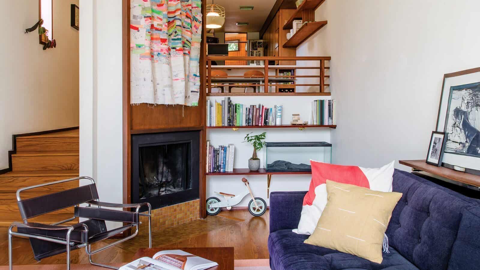 EmilyKeeney-officeofordinary_1600x900 Cozy living room featuring a purple sofa, modern chair, and fireplace. Shelves display books and a small bike. Stairs lead to a dining area partially visible above. Decorative art and pillows add charm to this space, reminiscent of Frank Lloyd-Wright's touch on home renovations by architects.