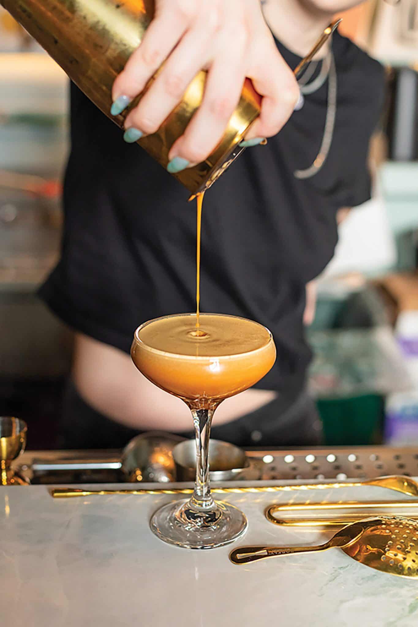 A person pours an espresso martini from a cocktail shaker into a coupe glass on a bar counter, capturing the vibe of new bars and restaurant openings in Seattle.