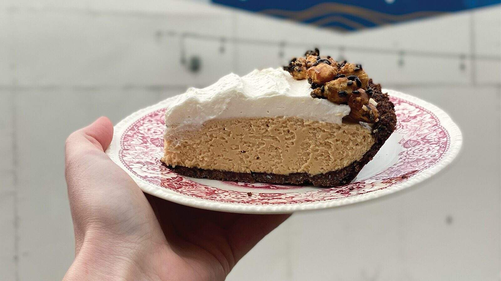 A hand holds a patterned plate with a slice of pie featuring a thick, creamy filling, a layer of whipped topping, and a crumbly crust with chocolate chips—perfect for those exploring where to eat in Seattle.